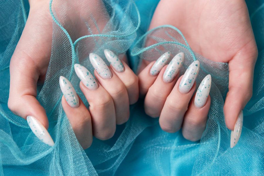 Hands with long almond-shaped nails in white polish with teal flakes on blue tulle fabric.