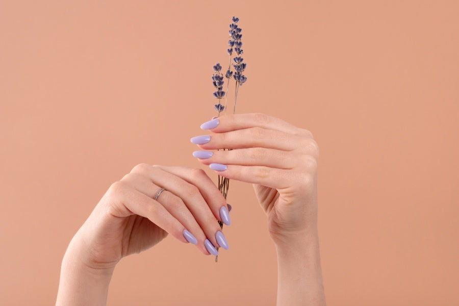 Hands with glossy lavender almond nails holding dried lavender stems on peach background.